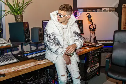 Un giovane skinny cantante con occhiali bianchi e giacca imbottita è seduto in uno studio di registrazione, circondato da tastiere e apparecchiature audio.