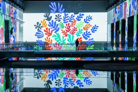 A lone figure stands on a walkway surrounded by vibrant Matisse cut-outs projected onto walls and reflected in the water at the Bassins des Lumières.