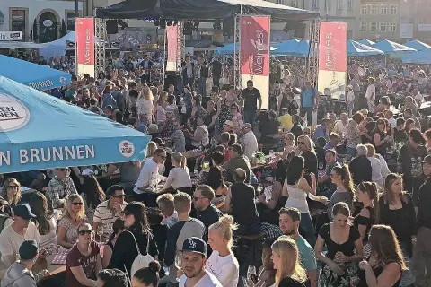 Menschenmenge feiert beim Bamberger Weinfest. Eine Bühne mit Musikern, Fahnen und Zelten. Viele Besucher sitzen an Tischen, trinken und genießen die Atmosphäre.