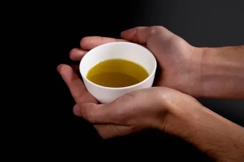 Mani che sorreggono una tazzina bianca con olio extravergine di oliva, evidenziando i suoi parametri visivi.