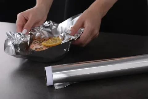 Mains de femme manipulant un saumon cuit dans du papier aluminium, prêt pour le nettoyage. Le rouleau d'aluminium est à côté.