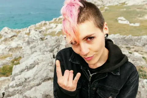 Dziewczyna z różowymi włosami i kolczykami, w kurtce dżinsowej, pozuje na tle skał i morza, pokazując znak rocka. Jej styl to czysty **punk**.