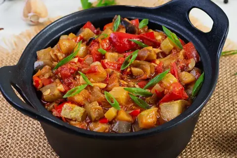Un plat de ratatouille colorée, avec des morceaux d'aubergine, de courgette, de poivron rouge et de tomate, garni de ciboule fraîche. Une recette savoureuse.