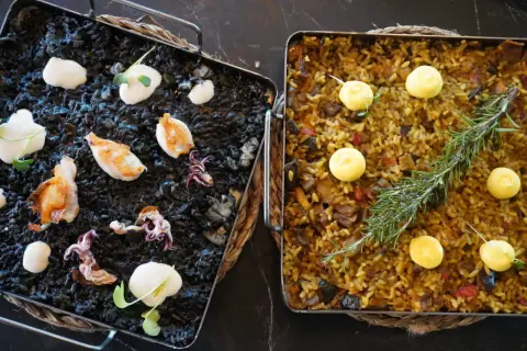 Dos arroces a la llauna: uno negro con calamares y alioli, y otro dorado con verduras y romero.
