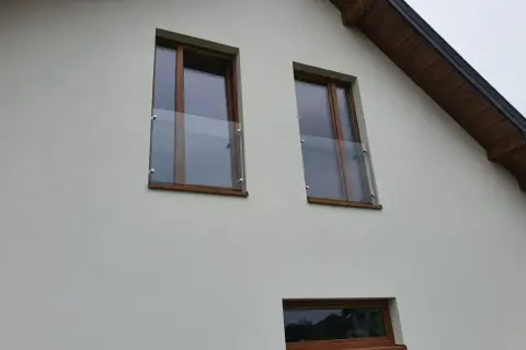 Nowoczesny portfenetr z drewnianymi ramami i szklaną balustradą przy oknach na białej elewacji.