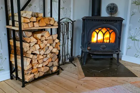 En mysig brasa sprakar i en gjutjärnskamin. Bredvid står en hylla fylld med ved, redo att installera braskamin för värme och trivsel.