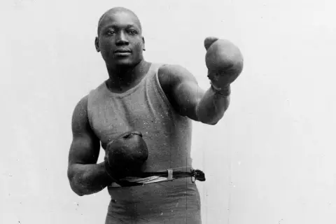 A boxer in an old boxing stance, wearing a tank top and shorts, with gloves on both hands, ready to fight.