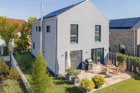 Ein modernes, schmales Haus mit Solaranlage auf dem Dach und einer Terrasse mit Grill und Sitzmöbeln.