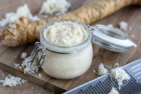 Frisch geriebener Meerrettich im Glas, bereit zur Haltbarmachung. Ein ganzer Meerrettich liegt daneben, daneben eine Reibe mit geriebenem Meerrettich.