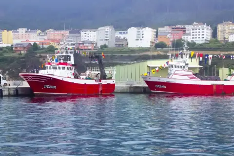 Dos barcos pesqueros rojos y blancos, listos para zarpar, adornados con banderas de gala. En el fondo, un pueblo costero con edificios de colores, evocando la tradición de la virxe da mariña.