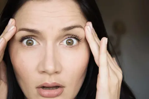 Mujer con ojos muy abiertos y arrugas en la frente, tocándose las sienes con las manos.