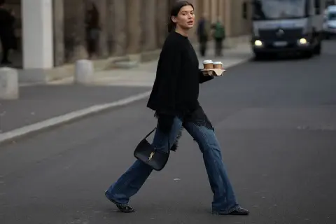 Kobieta w jeansach bootcut i czarnym swetrze z koronkowymi wstawkami niesie kawę. Stylowe jeansy bootcut to podstawa wielu casualowych stylizacji.