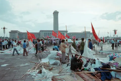 Ludzie na placu Tiananmen, czerwone flagi powiewają, budynki w tle, chaos i prowizoryczne namioty.