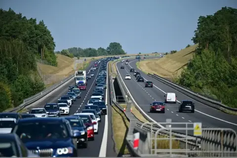Długi korek samochodów na autostradzie A6. Wiele pojazdów stoi w powolnym ruchu, otoczonych zielenią drzew.