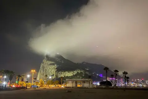 Nocny widok na Gibraltar, z chmurami otulającymi skałę. W oddali widać oświetlone miasto i palmy.