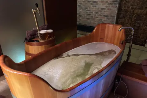 Entspannendes Bier-Wellness-Erlebnis in Tschechien: Ein hölzerner Bottich gefüllt mit schäumendem Bier, daneben ein Zapfhahn und zwei Biergläser.