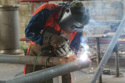 Spawacz w masce i rękawicach łączy metalowe rury. To praca, do której wykorzystuje się spawarkę inwertorową, generującą jasne iskry.