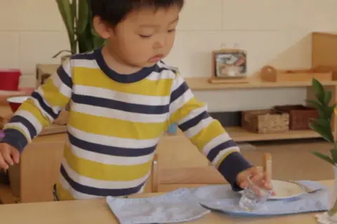 A young child, around 18 months old, practices setting a table in a Montessori environment, carefully placing a glass.