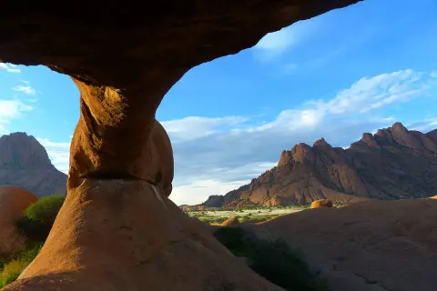 Felsformationen der Spitzkoppe in Namibia, mit einem Felsentor im Vordergrund und zerklüfteten Bergen am Horizont unter blauem Himmel.