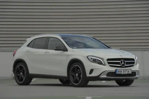 Biały Mercedes GLA na tle szarej, falistej ściany. Zbliżenie na przód auta, widoczne logo i tablica rejestracyjna. Idealny do miasta, poznaj jego mercedes gla wymiary.