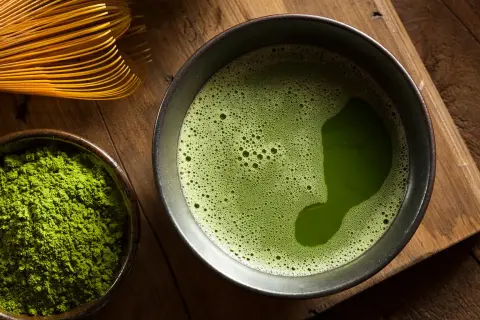 Une composition de matcha : poudre verte vibrante dans un bol, une tasse de thé mousseux et un fouet en bambou sur fond de bois.