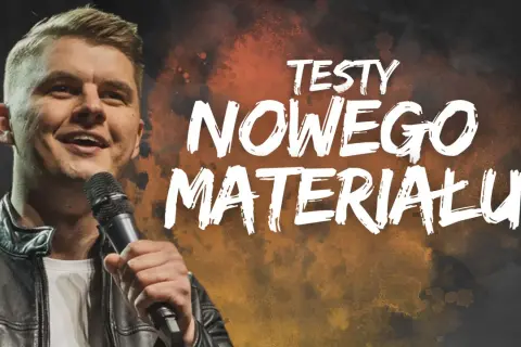 Uśmiechnięty komik na scenie, trzymający mikrofon. Na tle napis "TESTY NOWEGO MATERIAŁU".