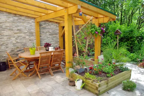 Un pergolato terrazzo fai da te accoglie un tavolo in legno e sedie, circondato da aiuole fiorite e vegetazione lussureggiante.