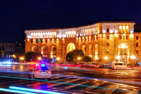Nocna panorama placu w Erywaniu, największym mieście Armenii. Oświetlony budynek z arkadami, na ulicy widać ślady świateł samochodów i radiowóz.