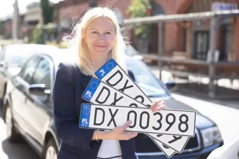 Kobieta trzyma tablice rejestracyjne z numerem DX 00398. Ułatwia to sd rejestracja.