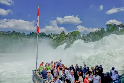 Menschen bestaunen den tosenden Rheinfall. Eine Schweizer Fahne weht im Wind. Der **Rheinfall Eintritt** ist ein beeindruckendes Naturschauspiel.