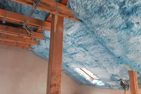 Piana na poddasze tworzy izolacyjną warstwę na drewnianej konstrukcji dachu, z widocznym oknem dachowym.