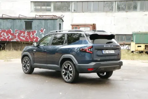 Ciemnoniebieska Dacia Bigster, model hybrydowy, zaparkowana na tle opuszczonej fabryki z wagonami kolejowymi pokrytymi graffiti.