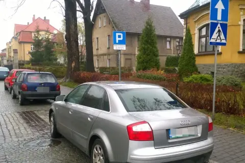 Samochody stoją na ulicy, a obok znak informuje o parkowaniu za przejściem dla pieszych.