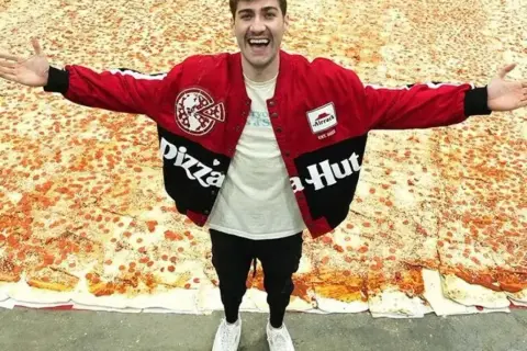 Mężczyzna w kurtce Pizza Hut stoi na gigantycznej pizzy.