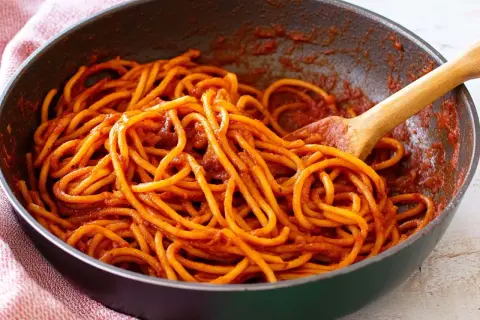 Spaghetti alla sorrentina in una padella, conditi con un ricco sugo rosso e mescolati con un cucchiaio di legno.