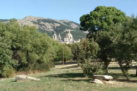 Un majestuoso edificio se alza entre los árboles del bosque de la Herrería, con montañas escarpadas al fondo bajo un cielo azul.