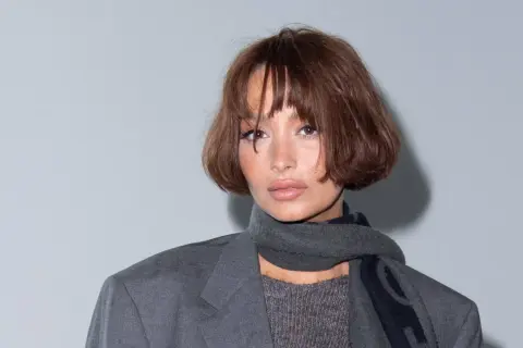 Frau mit braunem, kurzem Bob mit Curtain Bangs, trägt grauen Blazer und Schal.