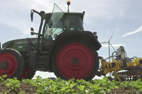Ein grüner Traktor mit roter Felge arbeitet auf einem Feld. Die Maschine ist mit einem gelben Anbaugerät ausgestattet, das zeigt, wie wird Reis geerntet.