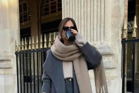 Stilvolle Frau trinkt Kaffee, kombiniert grau und beige mit einem Oversize-Schal und Mantel.