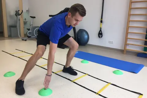 Mężczyzna w niebieskiej koszulce ćwiczy z drabinką koordynacyjną, zielonymi pachołkami i matą. W tle rower stacjonarny i drabinki.