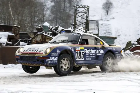 Porsche 959 w barwach Rothmans, z numerem 185, pokonuje śnieżny odcinek rajdu, wzbijając tumany śniegu.