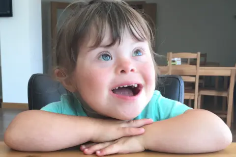 Una bambina down con occhi azzurri e un sorriso radioso, appoggiata al tavolo con le braccia incrociate.