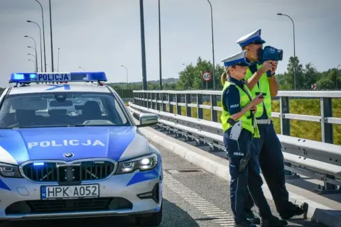 Policjanci kontrolują prędkość na drodze. Obok radiowóz z włączonymi sygnałami. Bezpieczne prowadzenie pojazdu to priorytet.
