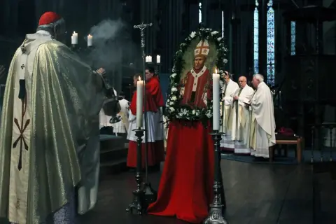 Ein Kardinal leitet eine gesungene katholische Messe. Ein Bildnis des Papstes, umrahmt von Blumen, steht im Mittelpunkt.