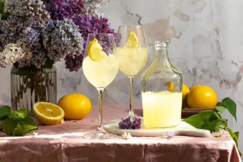 Zwei Gläser Limoncello Spritz mit Zitrone, eine Flasche Limoncello, Zitronen und Flieder auf rosafarbenem Tischtuch.