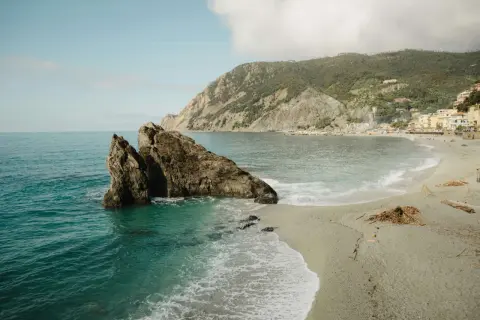 Un rocher émerge des eaux turquoise d'une crique. Une plage de galets s'étend vers un village coloré niché sur une colline verdoyante. Idéal pour savoir où partir en vacances cet été.