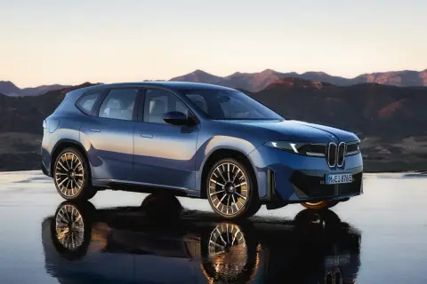 Niebieski BMW Neue Klasse, SUV z futurystycznym designem, stoi na mokrej nawierzchni z odbiciem gór w tle.