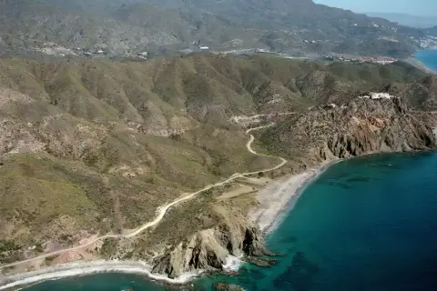 Vista aérea de la playa el lance, con su arena dorada, aguas turquesas y acantilados rocosos. Un camino serpentea por las colinas áridas.
