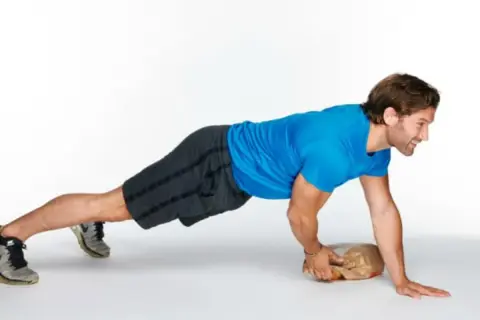 Man performing an around the world plank exercise, balancing on one hand and a paper bag.