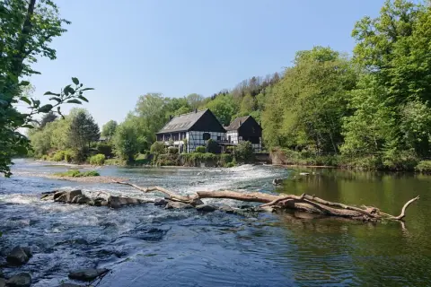Fachwerkhäuser am Flussufer, wo die **Wipperkotten wandern**. Ein Baumstamm liegt im rauschenden Wasser.